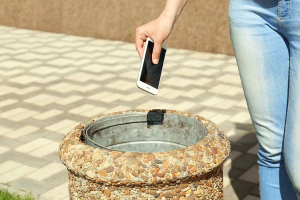 Young woman throwing smartphone in litter bin outdoors