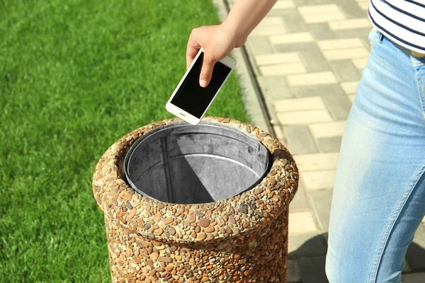 Young woman throwing smartphone in litter bin outdoors