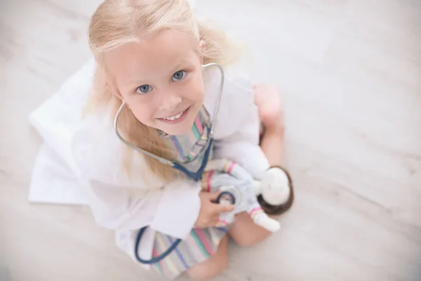 Menina em casaco médico brincando com boneca no chão — Fotografia de Stock