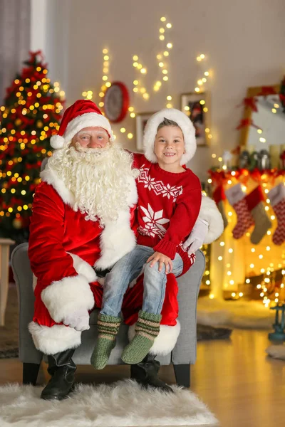 Glad liten pojke som sitter i tomtens knä i rum med vackra juldekorationer — Stockfoto