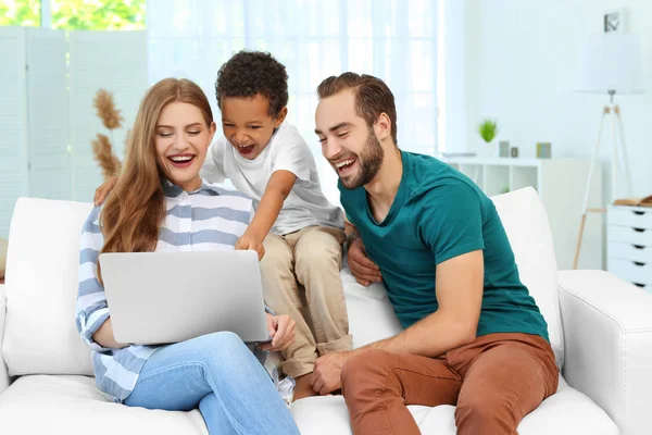 Gelukkige paar met goedgekeurde Afro-Amerikaanse jongen met behulp van laptop zittend op de Bank thuis — Stockfoto