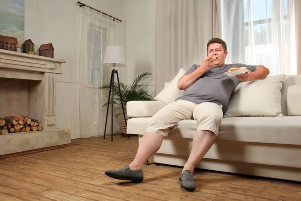 Túlsúlyos fiatal ember eszik édességet a kanapén, otthon — Stock Fotó