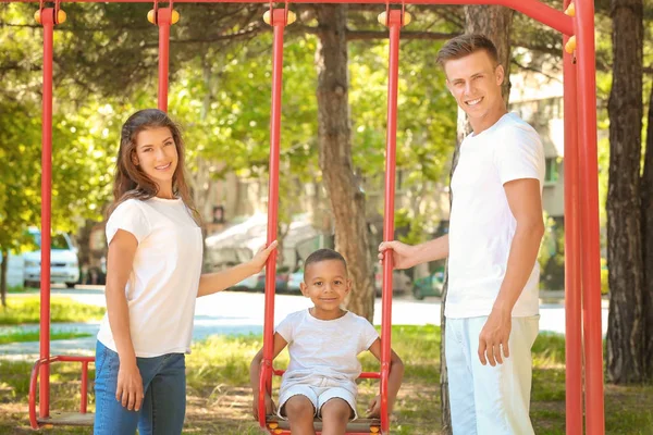 Jovem família com adotivo menino afro-americano jogando em balanços no parque — Fotografia de Stock