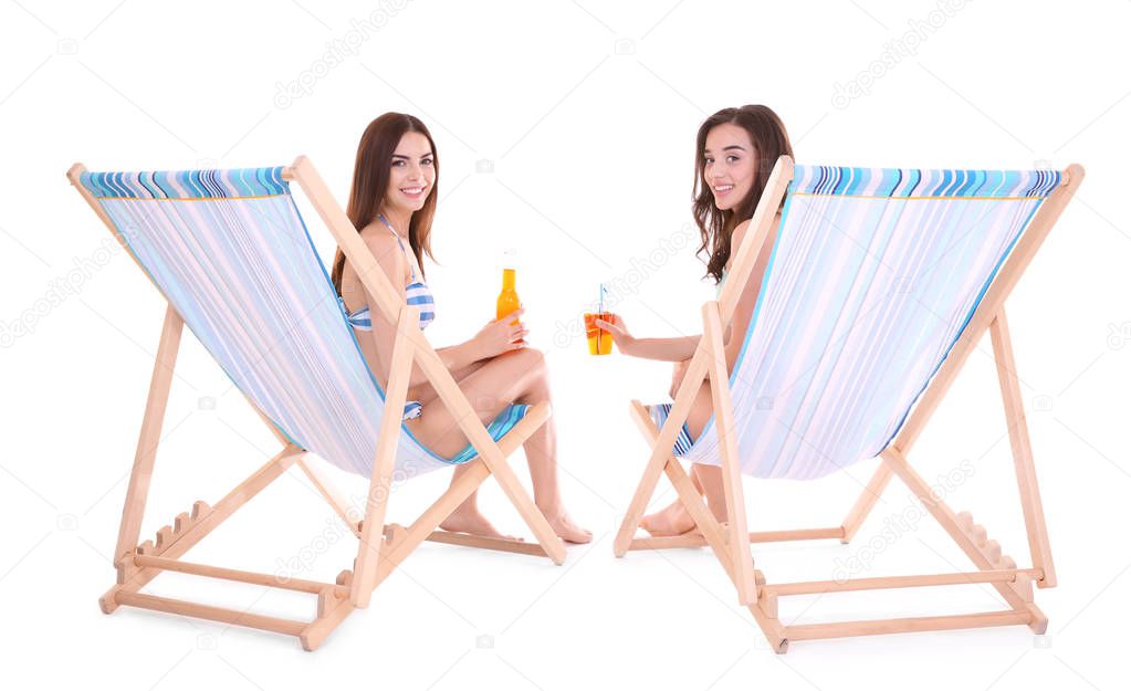 Beautiful young women with alcoholic drinks sitting on deck chairs against white background