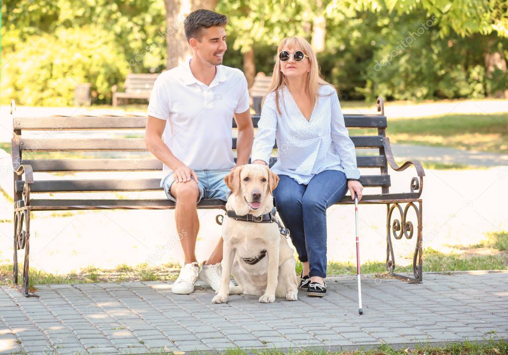 Young man and blind woman with guide dog sitting on bench in park