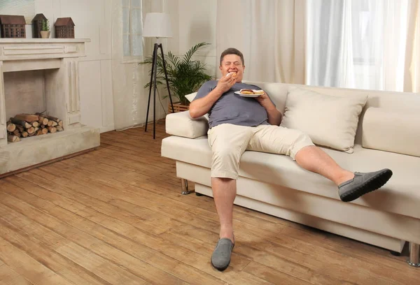 Надмірна вага молодий чоловік їсть цукерки на дивані вдома — стокове фото