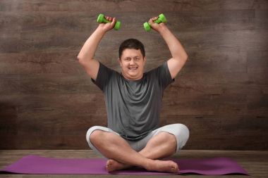 Overweight man training with dumbbells against wooden wall clipart