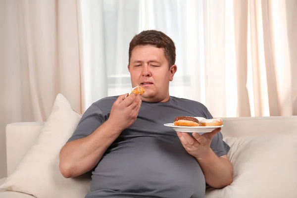 Młody mężczyzna jedzenie słodyczy na kanapie w domu z nadwagą — Zdjęcie stockowe