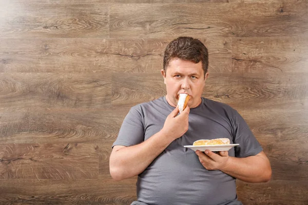 Overweight young man eating sweets on wooden background — Stock Photo, Image