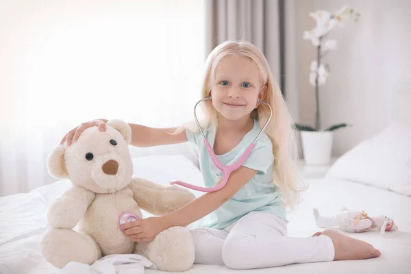 Little girl playing with teddy bear and stethoscope on bed — Stock Photo, Image