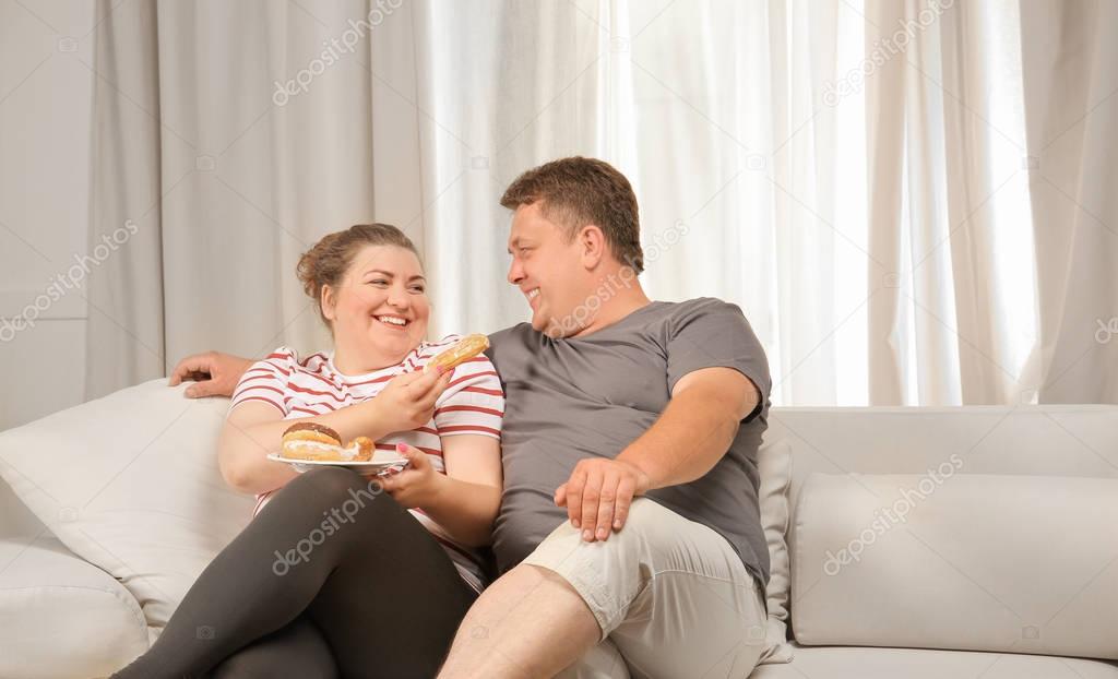Overweight couple eating sweets on sofa at home
