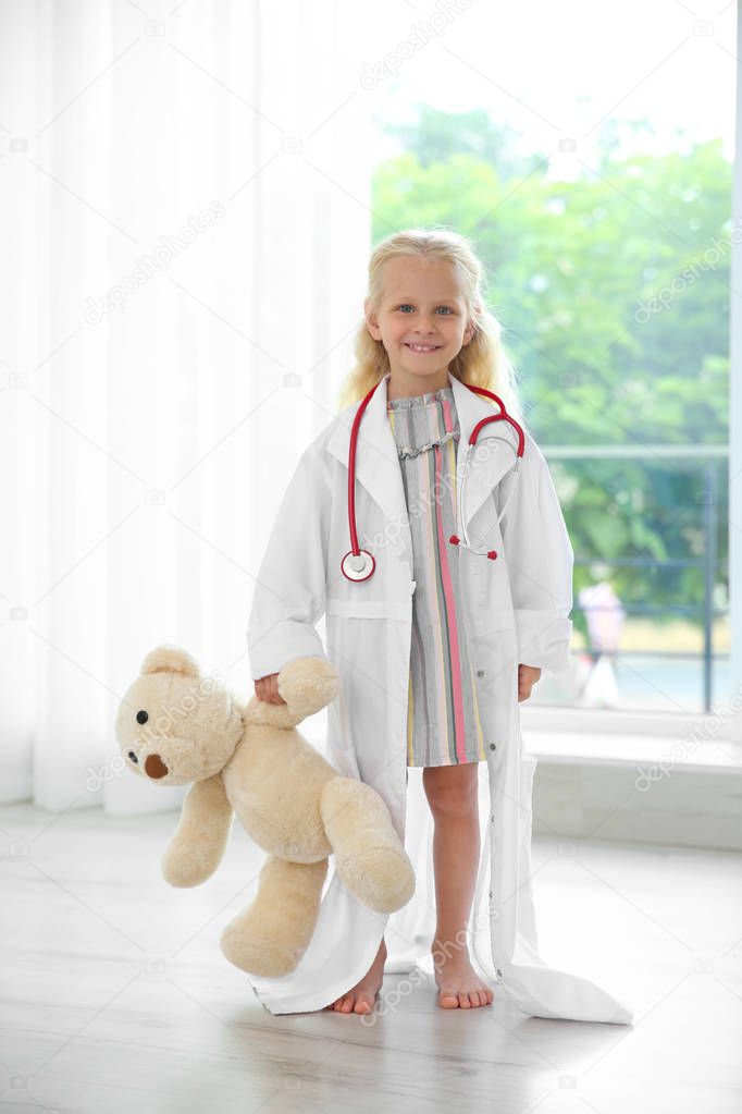 Little girl in medical coat with teddy bear standing near window