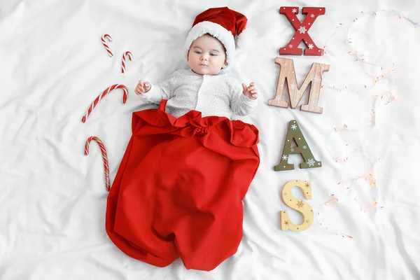 Bebê bonito usando chapéu de Papai Noel em saco vermelho e palavra XMAS feito de letras de madeira com luzes de Natal em folha branca — Fotografia de Stock
