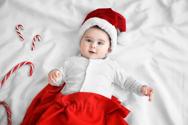 Niedliches kleines Baby mit Weihnachtsmütze in roter Tüte und Bonbons auf weißem Laken — Stockfoto