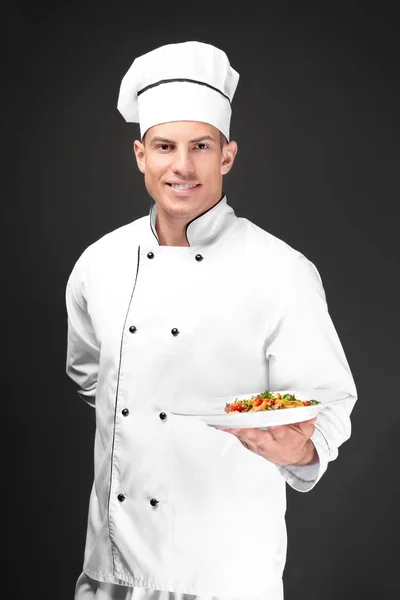 Young male chef holding plate with salad on dark background — Stock Photo, Image