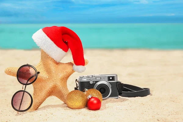 Chapéu de Santa com estrela do mar, câmera e óculos de sol na praia. Natal feriados conceito — Fotografia de Stock