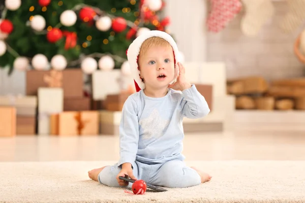 Niedliches kleines Baby mit Weihnachtsmütze im weihnachtlich dekorierten Zimmer — Stockfoto