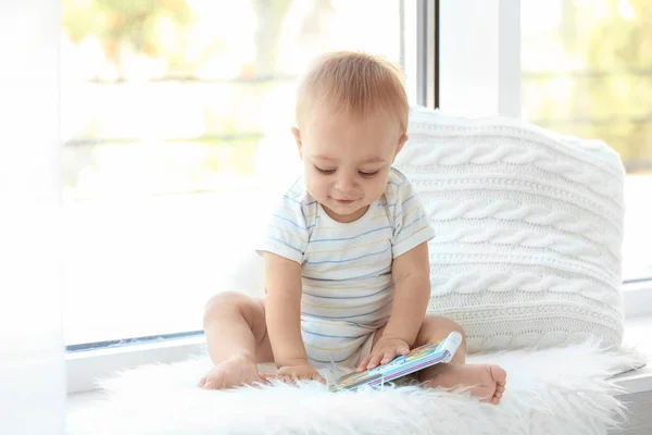 Cute little baby with book sitting on window sill at home — Stock Photo, Image