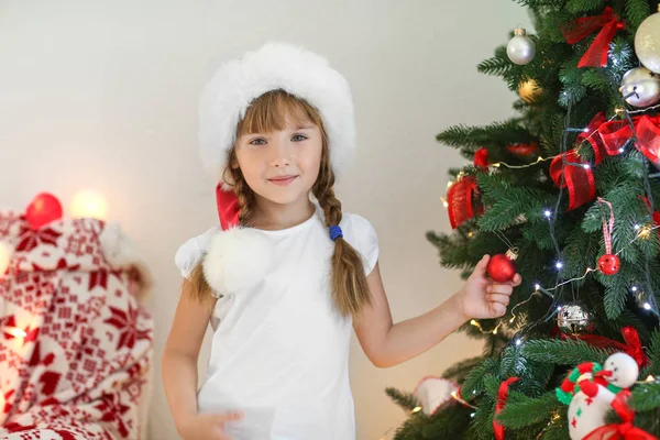 可爱的小女孩在圣诞老人的帽子装饰圣诞树在家里 — 图库照片