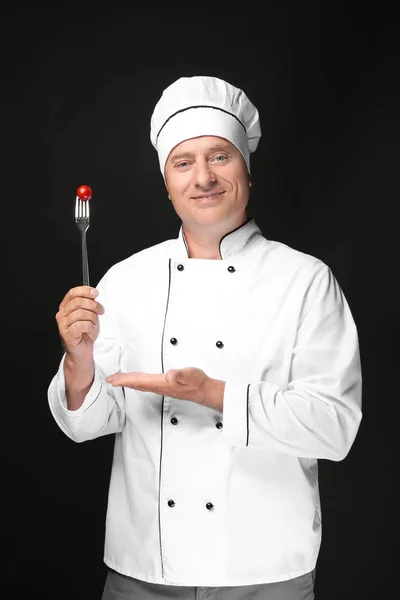 Male chef in uniform holding fork with cherry tomato on black background — Stock Photo, Image