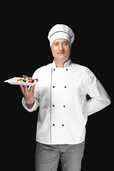 Manlig kock i uniform håller plattan med sallad på svart bakgrund — Stockfoto