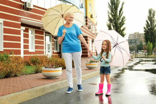 Starší žena a malá holčička chůzi na ulici s deštníky — Stock fotografie