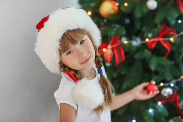 可爱的小女孩在圣诞老人的帽子装饰圣诞树在家里 — 图库照片