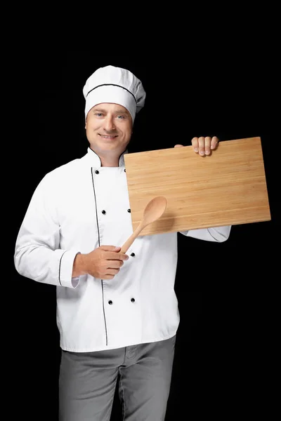 Male chef in uniform holding wooden spoon and board on black background Stock Picture