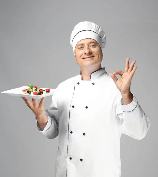 Чоловічий шеф-кухар в уніформі тримає тарілку з салатом на сірому фоні — стокове фото