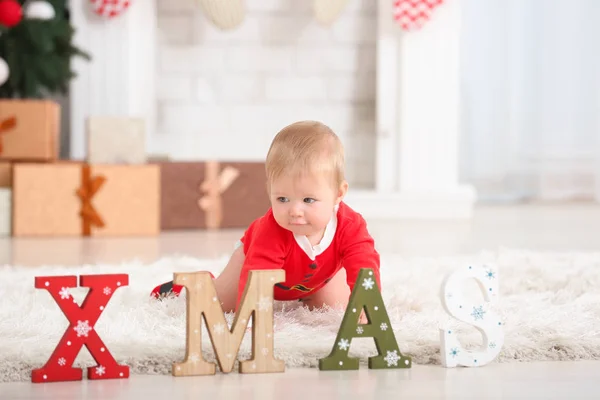 Χαριτωμένο μωρό στο κοστούμι του Άγιου Βασίλη και λέξη Χριστούγεννα στο πάτωμα στο σπίτι — Φωτογραφία Αρχείου