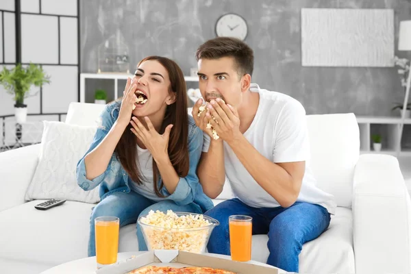 Jovem casal comer pipocas enquanto assiste TV no sofá em casa — Fotografia de Stock