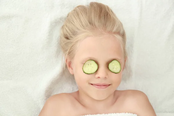 Piccola ragazza carina con fette di cetriolo su occhi sdraiati su tessuto leggero — Foto Stock
