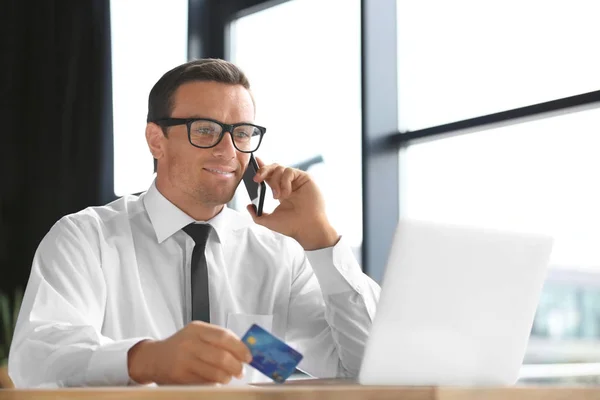 Jovem empresário segurando cartão de crédito enquanto faz chamada no local de trabalho — Fotografia de Stock