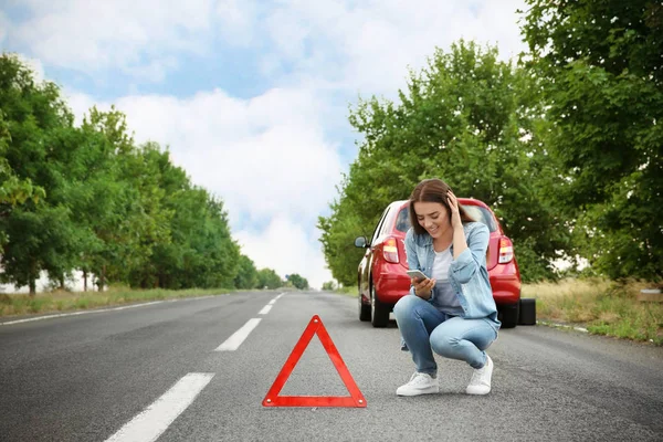 Jonge vrouw met mobiele telefoon zitten op weg in de buurt van gebroken auto boos — Stockfoto