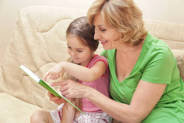 Linda chica con la abuela leyendo libro en el sofá en casa — Foto de Stock