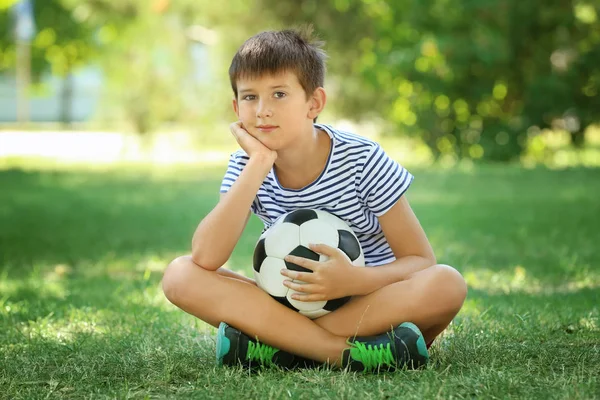 Rapaz bonito com bola de futebol sentado no gramado no parque — Fotografia de Stock