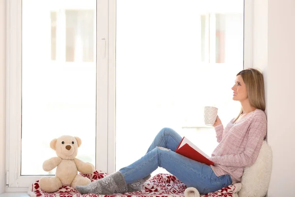 Joven hermosa mujer con libro y taza de bebida sentado en el alféizar de la ventana. concepto de vacaciones de Navidad — Foto de Stock