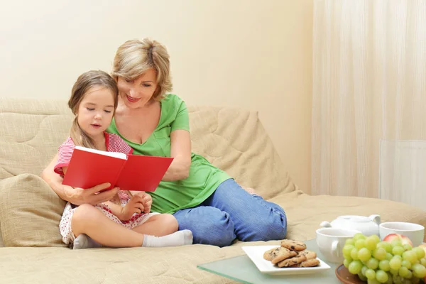Милая девушка с бабушкой читает книгу на диване дома — стоковое фото