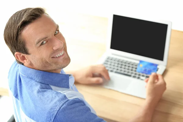 Junger Mann mit Kreditkarte, während er Laptop auf hellem Hintergrund benutzt — Stockfoto