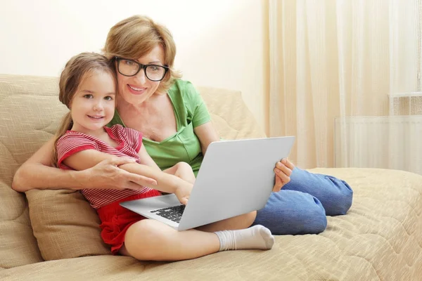 Cute girl and grandmother with laptop on sofa at home — Stock Photo, Image