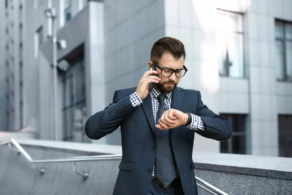 Hombre de negocios guapo hablando por teléfono móvil mientras mira el reloj al aire libre — Foto de Stock