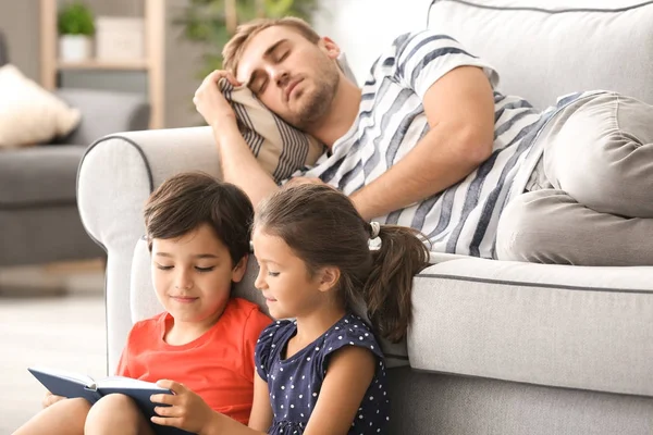 Lindos niños leyendo libro mientras su padre está durmiendo en el sofá en casa —  Fotos de Stock