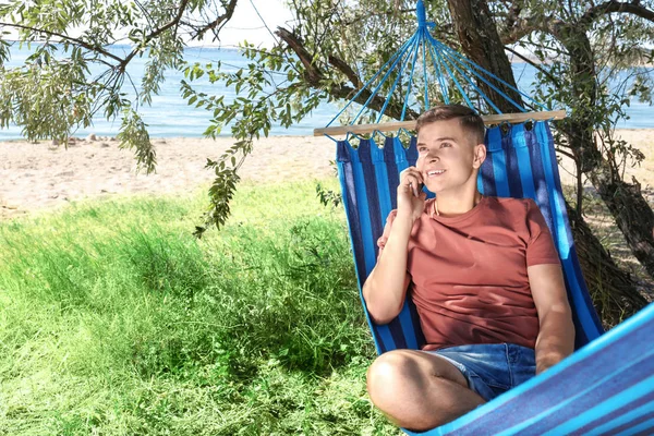 Jovem falando por telefone celular enquanto descansa na rede ao ar livre — Fotografia de Stock