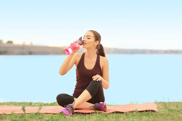 Jonge sportieve vrouw zitten en drinken van koud water op de kust rond de rivier — Stockfoto