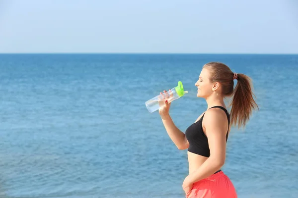 Jovem mulher esportiva com garrafa de água na praia do mar — Fotografia de Stock