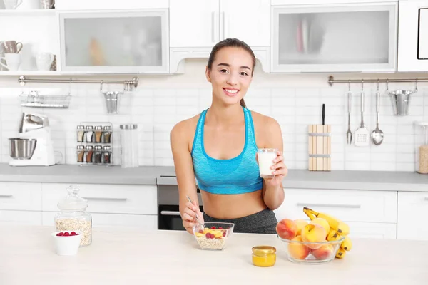 Молодая женщина в спортивной одежде пьет молоко и ест овсянку на кухне — стоковое фото