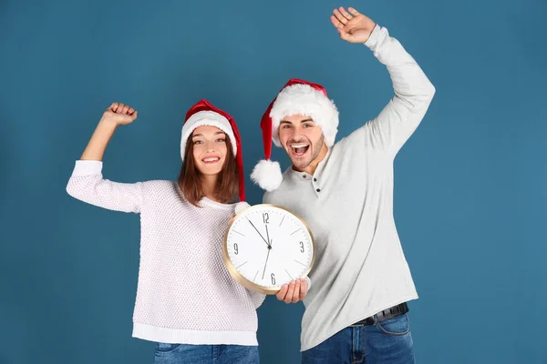 年轻夫妇在圣诞老人帽子与时钟在颜色背景。圣诞倒计时概念 — 图库照片