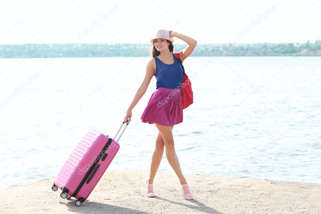 Beautiful young tourist with suitcase standing on river bank