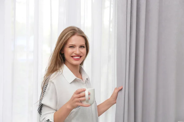 Młoda kobieta z filiżankę kawy stojący w pobliżu zasłony w domu — Zdjęcie stockowe