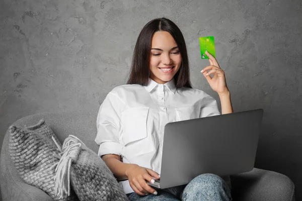 Mujer joven usando el ordenador portátil mientras sostiene la tarjeta de crédito sobre fondo gris. Concepto de compras por Internet — Foto de Stock